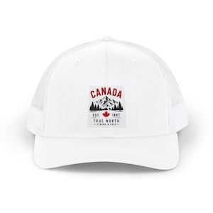 May include: A white trucker cap featuring a graphic print on the front panel. The design includes the red text CANADA above a black mountain and forest illustration, a red maple leaf, and the text EST. 1867 TRUE NORTH - STRONG & FREE -.