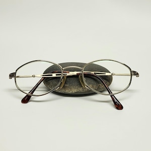 May include: Oval-shaped spectacles with a thin, metallic gold-tone frame. The temples feature a dark brown tortoiseshell pattern, and the bridge has a delicate, open-work design. These spectacles have clear lenses and adjustable nose pads.