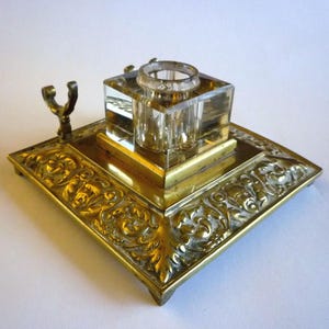 May include: A square brass inkwell stand featuring an ornate embossed floral border and a small metal pen rest on one side. A clear, thick-walled glass ink reservoir sits centred on the metallic base, which is supported by four small feet.