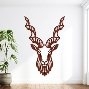 May include: A dark brown wooden wall art piece featuring a stylised, symmetrical cutout design of a markhor goat head. The artwork displays the animal's distinct long, spiralling horns with chevron patterns and a detailed face with a beard.