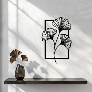 May include: A black metal wall art piece featuring a rectangular frame containing three ginkgo biloba leaves with detailed vein patterns. The leaves are connected by thin stems that extend from the bottom of the frame, creating a botanical design.