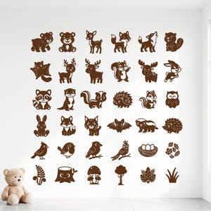 May include: A collection of brown wall decals featuring various woodland animals, birds, plants, and nature elements arranged in a grid pattern on a white wall, accompanied by a plush teddy bear sitting on the floor in the lower left corner.