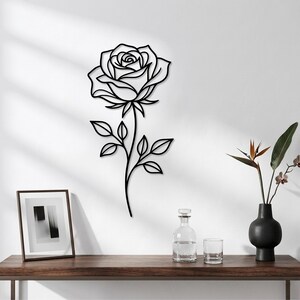 Può includere: Un'opera d'arte da parete in metallo nero a forma di rosa singola con un lungo stelo e quattro foglie. Il design presenta linee pulite e continue che formano i petali e il fogliame, creando una decorazione floreale piatta da parete.