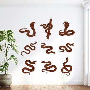 May include: A collection of nine dark brown wooden wall art pieces shaped like various snakes, including a cobra, a serpent coiled around a staff, and several winding snakes in different poses, arranged in a grid pattern on a white wall.
