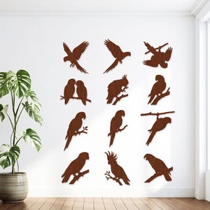 May include: A collection of twelve dark brown wooden wall art pieces, each shaped as a silhouette of a parrot or cockatoo in various poses, including birds in flight, perched on branches, or preening, arranged in a grid on a white wall.