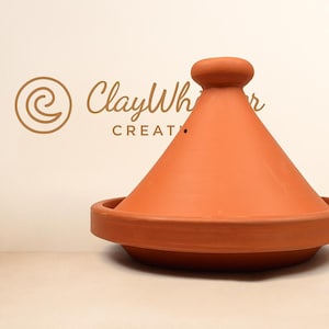Op de afbeelding: Een terracotta tajine-pot met een conisch deksel en een ronde basis. De pot heeft een warme oranje kleur en staat op een licht oppervlak. Het logo "ClayWhisper Creations" is zichtbaar op de achtergrond.