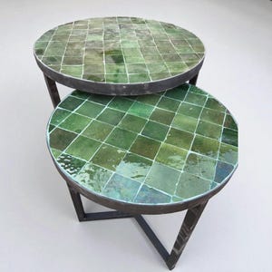 May include: Two round side tables with green tiled tops and dark metal frames. The tables are stacked, with the top table slightly smaller than the bottom one. The tiled surfaces are a mosaic of green squares.