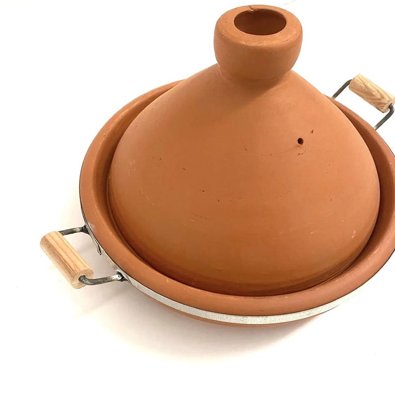 May include: A terracotta tagine pot with a conical lid and a wide base. The pot has a natural, earthy color and features metal handles with wooden grips. The base has a white stripe around the rim.