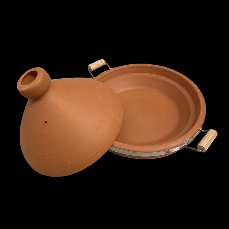 May include: A terracotta tagine pot with a matching lid, set against a black background. The lid features a conical top with a vent. The pot has a metal rim and wooden handles. A traditional cooking vessel.