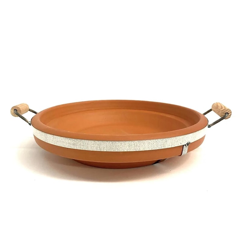 May include: A terracotta-colored, shallow cooking dish with a white band around the middle. It has two handles with wooden grips and metal supports. The dish is designed for cooking and serving.