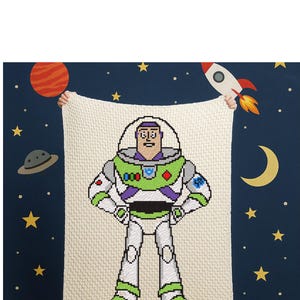Könnte beinhalten: Eine cremefarbene, pixelige Decke mit Buzz Lightyear aus Toy Story. Die Decke hat ein grünes, weißes und lila Design, mit einem Weltraum-Hintergrund aus Sternen, Planeten und einer Rakete.