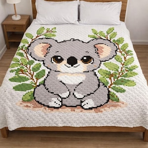 Puede incluir: Una manta de ganchillo con un diseño de koala gris, pecho blanco y orejas beige, rodeado de ramas verdes. El koala está sobre un fondo blanco. La manta se muestra en una cama de madera con almohadas blancas.