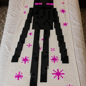 May include: A white crocheted blanket with a black pixelated figure and pink eyes, plus pink star and cross shapes. The blanket is rectangular and lies on a bed.