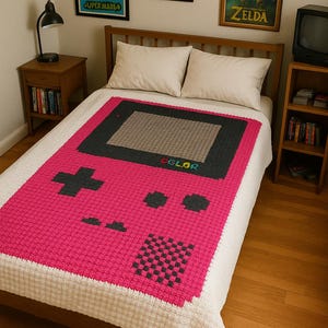 May include: A pink, gray, and black crocheted blanket designed to resemble a retro handheld video game console. The blanket features a cross-shaped directional pad, buttons, and a screen. The word "COLOR" is visible on the blanket. The blanket is on a bed with framed artwork.