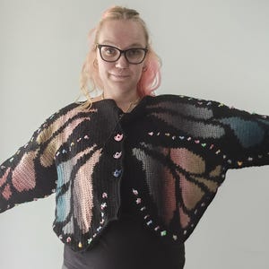 Puede incluir: Un cárdigan de ganchillo negro con un patrón de alas de mariposa en tonos rosa, azul y beige. La prenda tiene mangas largas y se abrocha con cuatro pequeños botones coloridos en forma de tetera en la parte delantera central.