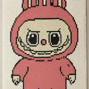 May include: A pixelated pink bunny character with a cream-colored face and black outlines. The bunny is wearing a pink hooded jumpsuit. The background is a cream-colored knitted fabric.