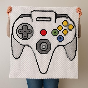 May include: A white blanket with a pixelated design of a video game controller. The controller has a grey directional pad, a red button, and colourful buttons. The blanket is held up by a person.