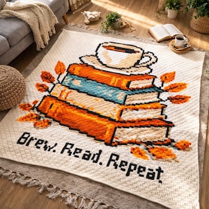 Puede incluir: Una manta de crochet color crema con un diseño pixelado que presenta una pila de libros, una taza de café y el texto "Brew. Read. Repeat". Los libros son naranjas y azules, con hojas naranjas. La manta está flecada.