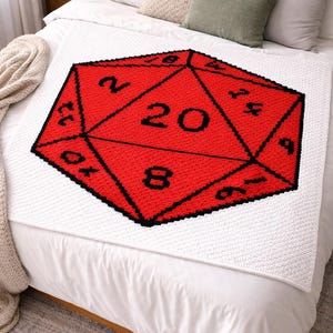 May include: A handmade, square, white blanket with a large red and black twenty-sided die design. The die features the number 20 prominently, with other numbers visible. The blanket is displayed on a bed with pillows.