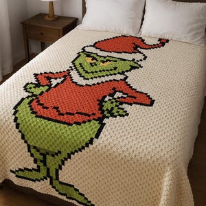 May include: A cream-coloured crocheted blanket depicting a pixelated green character in a red Santa hat and sweater. The blanket is on a bed with white pillows.