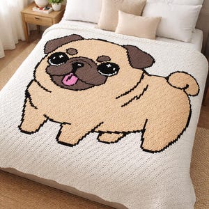Puede incluir: Una manta de crochet color crema con un gran diseño de perro pug pixelado. El pug tiene pelaje marrón claro, orejas marrones y una lengua rosa. La manta está sobre una cama con almohadas blancas y un cabecero neutro.