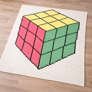 Puede incluir: Una alfombra de ganchillo con un diseño de Cubo de Rubik pixelado. El cubo está representado en colores amarillo, rojo y verde, con contornos negros. La alfombra tiene un fondo blanco roto neutro y está colocada sobre un suelo de madera.