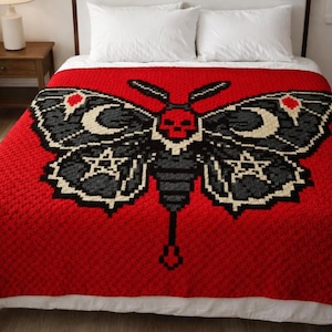 Könnte beinhalten: Eine rote Häkeldecke mit einem großen Mottenmuster. Die Motte hat schwarze und graue Flügel mit einem Totenkopf, Monden und Sterndetails. Die Decke liegt auf einem Bett mit weißen Kissen.