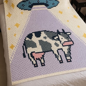 Puede incluir: Manta de ganchillo con diseño pixelado. Presenta un OVNI azul y gris sobre un rayo púrpura, y una vaca en blanco y negro. El fondo es crema con acentos de estrellas amarillas.