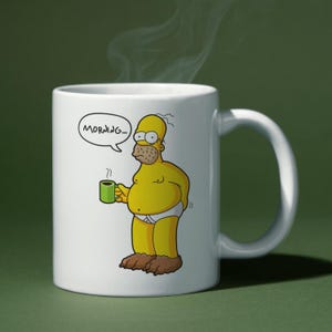May include: White ceramic mug featuring a cartoon image of Homer Simpson in his underwear, holding a green mug of coffee. A speech bubble above his head says "MORNING...". The mug has a white handle and is set against a green background.