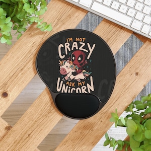 Puede incluir: Alfombrilla de ratón ergonómica negra con reposamuñecas. La alfombrilla presenta una imagen de dibujos animados de Deadpool montando un unicornio, con el texto "I'M NOT CRAZY ASK MY UNICORN" en blanco y rojo.