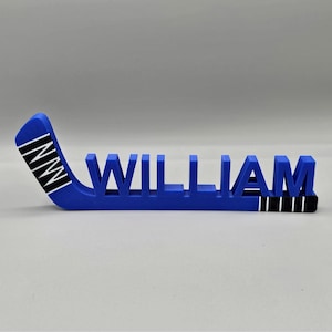 May include: A blue hockey stick-shaped nameplate with the name "WILLIAM" in capital letters. The hockey stick blade has the letters "INNI" in white. The base of the nameplate has black and white stripes.