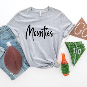 May include: A gray t-shirt with the word "Mounties" written in black cursive. The shirt is laid out on a white surface with a football, jeans, a beer bottle, and a green and white pennant.