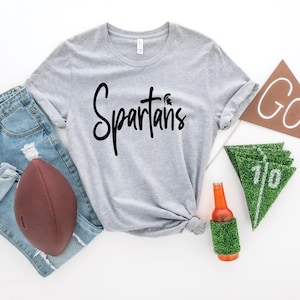 May include: A gray t-shirt with the text "Spartans" in black lettering. The shirt is laid out on a white surface with a football, jeans, a beer bottle, and pennants.