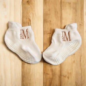 May include: A pair of white baby socks with a non-slip grip sole. Each sock has the embroidered initials "BM" in a copper color. The socks are displayed on a light brown wooden surface.