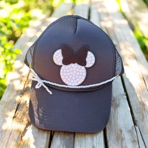 May include: Black trucker hat with a Minnie Mouse design. The design features a black bow and white mouse ears filled with iridescent rhinestones. A white beaded string accents the brim, tied with a bow.
