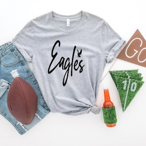 May include: A gray t-shirt with the word "Eagles" in black script with a small black bird graphic above the word. The shirt is laid out on a white surface with a football, jeans, a brown pennant with the word "GO!" on it, and a green and white pennant with the number "110" on it.