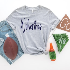 Op de afbeelding: Grijs T-shirt met de tekst "Wolverines" in donkerblauwe schrijfletters met een pootprint boven de tekst. Het shirt ligt op een wit oppervlak met een voetbal, jeans, een bierfles en vlaggetjes.