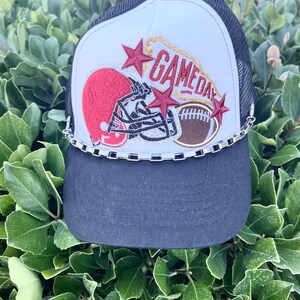 May include: White and black trucker hat with a red, white, and gold football helmet and football design. The hat has a silver chain trim and the words "GAMEDAY" embroidered on the front.
