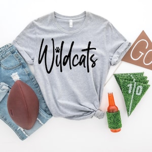 May include: Gray t-shirt with the text "Wildcats" in black with a paw print above the word "Wildcats". The shirt is laid out on a white surface with a football, jeans, a beer bottle in a grass koozie, and pennants that say "GO!" and "10".