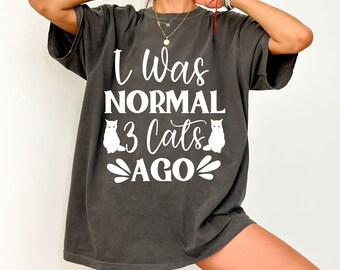 May include: Dark grey t-shirt with the text "I Was NORMAL 3 Cats AGO" in white script. Two white cat illustrations are on the shirt. The shirt is a casual, oversized style.
