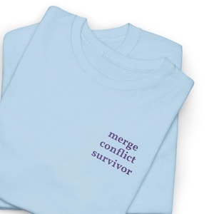 Puede incluir: Dos camisetas de color azul claro apiladas una encima de la otra. El texto "merge conflict survivor" está impreso en morado en la parte superior izquierda del pecho de la camiseta superior. Las camisetas están dobladas y parecen ser de material suave.