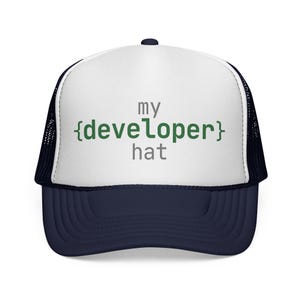May include: A navy blue and white trucker hat with the text "my {developer} hat" in gray and green. The hat has a mesh back and a curved brim.
