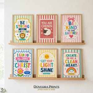 May include: Six framed wall art prints with light-colored wooden frames, arranged on two shelves. Each print features colorful, whimsical illustrations and inspirational text, including phrases like Be Strong, You Are Chosen, and Let Your Light Shine.