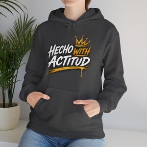 May include: Dark gray hoodie with a front pocket and drawstring hood. The hoodie features the text "HECHO WITH ACTITUD" in white and gold, with a gold crown graphic above the text. The design has a distressed look.