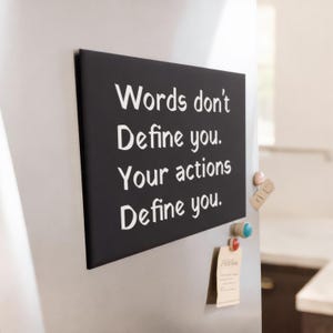 May include: A black rectangular sign with white text that reads "Words don't Define you. Your actions Define you." The sign is attached to a stainless steel refrigerator door with magnets. Other items are attached to the refrigerator.