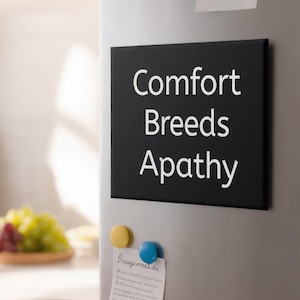 May include: A black rectangular sign with the words "Comfort Breeds Apathy" in white lettering is attached to a refrigerator. Two magnets, one yellow and one blue, are below the sign. A handwritten note is attached with the blue magnet.