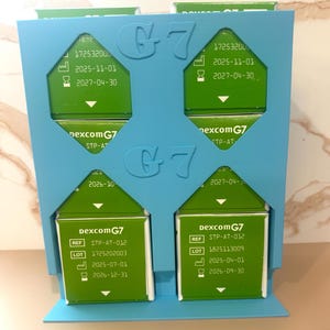 May include: A blue holder with four green Dexcom G7 boxes. Each box has white text with the product name, reference number, lot number, and expiration dates. The holder has the letters "G7" in a raised design.