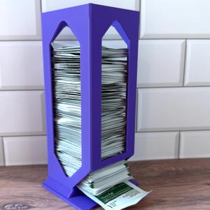 May include: A vertical purple plastic dispenser with a rectangular frame and open sides, filled with a stack of white square packets. The bottom packet is partially pulled out, showing the text Thick Pad Offers Excellent Cleansing Action.