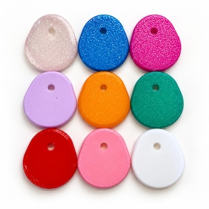 Puede incluir: Una colección de nueve colgantes en forma de lágrima dispuestos en una cuadrícula de tres por tres. Cada colgante presenta un único orificio centrado y viene en un color distinto, incluyendo blanco brillante, azul, rosa, morado, naranja, verde, rojo, rosa claro y blanco sólido.