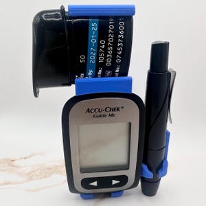 May include: An Accu-Chek Guide Me blood glucose meter with a black and silver design, a black test strip container, and a black lancing device. The meter has a digital display and blue plastic holders.